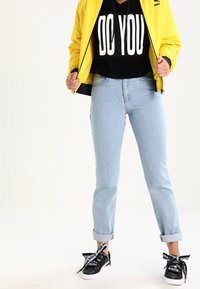 Gelbe gesteppte Jacke über einem schwarzen Hoodie mit "DO YOU" Aufdruck, hellblaue hochgekrempelte Jeans und schwarze Sneaker mit Logo-Schnürsenkeln.