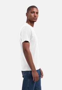 Weißes T-Shirt mit kurzen Ärmeln aus Baumwolle, Rundhalsausschnitt und geradem Saum. Das Modell trägt blaue Jeans und steht leicht zur Seite.