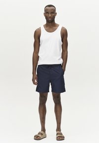 Marineblaue Badeshorts mit Kordelzug, kombiniert mit einem weißen Tanktop und hellbraunen Sandalen. Glattes Material, lässiges Design, knielange Shorts.