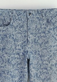 Pantalons en denim bleu avec un motif en dentelle florale blanche, comportant une poche avant et une fermeture à bouton métallique à la taille.