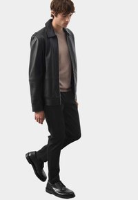 Czarna skórzana kurtka o gładkiej fakturze, z płaskim kołnierzem i zamkiem błyskawicznym z przodu, w zestawieniu z beżowym swetrem i czarnymi spodniami. Czarne buty dopełniają wygląd.