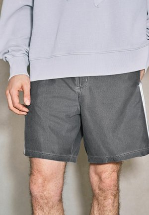 Bărbat poartă pantaloni scurți gri și o cămașă cu mâneci lungi gri deschis, arătând mâinile și picioarele pe un fundal neutru.