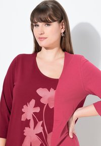 Maroones Oberteil mit floralem Design in hellrosa. Hat einen runden Ausschnitt und Dreiviertelärmel. Weiche, glatte Stofftextur.