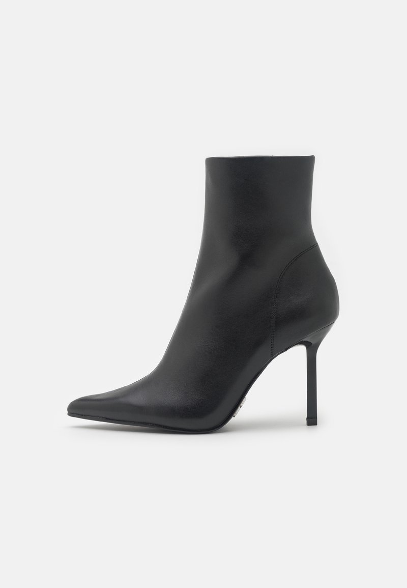 Bottine en cuir noir à bout pointu et talon stiletto haut et fin sur un fond gris clair uni.