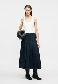 Γυναίκα που φοράει λευκό τοπ χωρίς μανίκια, σκούρα πλισέ μίντι φούστα, μαύρες μποτάκια, κρατώντας μια μαύρη δερμάτινη τσάντα με αλυσίδα λεπτομέρειας.