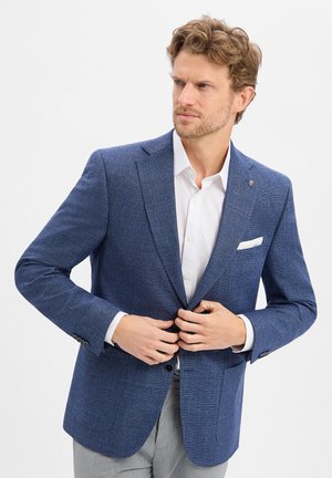 Mann mit kurzem lockigem Haar, der einen blauen strukturierten Blazer über einem weißen Hemd zuknöpft, trägt hellgraue Hose und schaut zur Seite.