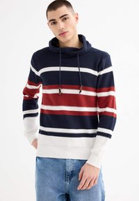 Joven vestido con suéter de punto texturizado con rayas horizontales en azul marino, rojo y blanco, combinado con jeans azul claro, de pie contra un fondo blanco.