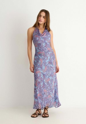 Femme portant une robe maxi sans manches à imprimé cachemire bleu et rose, avec un décolleté drapé et des sandales plates marron à brides.