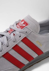 Grå mockaskor med röda randiga detaljer, med en texturerad yta, runda snören och guld "JEANS"-detalj på sidan.