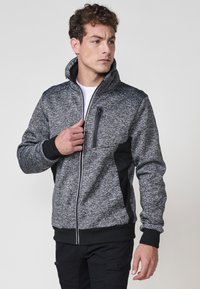 Chaqueta de tejido texturizado en gris y negro, con cierre de cremallera, cuello alto, bolsillos laterales y un bolsillo en el pecho. Presenta puños y dobladillo acanalados.