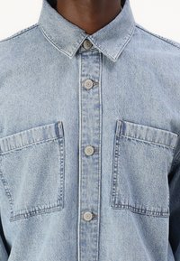 Ανοιχτό γαλάζιο τζιν πουκάμισο με αιχμηρό γιακά, κεντρική κουμπωμένη στερέωση και δύο τσέπες στο στήθος με αντίθεση ραφή και μεταλλικά εξαρτήματα.
