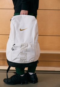 Bijeli Nike ruksak sa zlatnim naglascima, s velikim natpisom "ELITE", patentnim zatvaračima i crnom bazom. Glatka tekstura s mrežastim bočnim džepom.