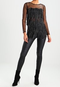 Top lungo in mesh nero con dettaglio frange, caratterizzato da un'impermeabile trasparente e un motivo a pois. Abbinato a leggings neri aderenti e stivaletti alla caviglia.