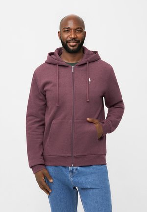 Bordeauxfarbene Zip-Hoodie mit einer Brusttasche, Kapuze mit Kordelzug und kleinem Baumlogo. Hat eine weiche Textur und einen lässigen Schnitt. Über einem grünen Shirt getragen.