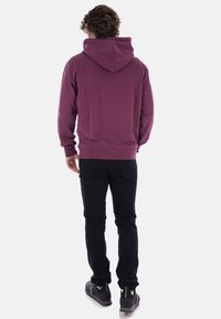 Sudadera con capucha color burdeos hecha de tela suave, con un bolsillo tipo canguro y puños acanalados. Se lleva con jeans negros y zapatillas.