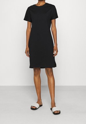 Personne portant une robe noire unie à manches courtes, longueur genoux, et des sandales blanches à enfiler, debout devant un fond blanc uni.