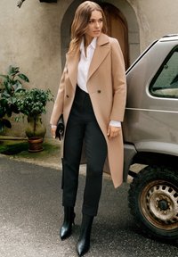 Długi beżowy wełniany płaszcz z dużymi klapami, w połączeniu z białą koszulą i czarnymi spodniami o wysokim stanie. Czarne botki na kostkę i mała kopertówka.