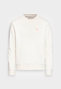 Sudadera de color crema hecha de tejido suave. Presenta un escote redondo, mangas largas, puños de canalé y un pequeño parche de logo rosa en la parte delantera.