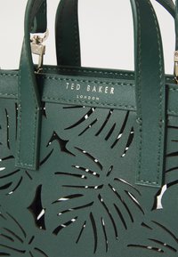 Zöld bőr kézitáska, kivágott levélmintázattal, arany színű "Ted Baker London" logóval és strapabíró felső fogantyúkkal.