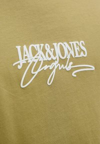 Olivu zaļa kokvilnas krekls ar izteiktu baltu tekstu "JACK & JONES Originals", kur burts "O" ir izcelt ar slīpo rakstu. Gluda tekstūra.