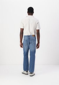 Muž v lehké košili s krátkým rukávem, modrých džínách a bílých teniskách stojí čelem ke bílé zdi, s uvolněnými rukama podél těla.