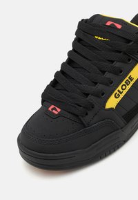 Crne skate tenisice s teksturiranom površinom, ukrašene žutim i crvenim detaljima. Debele crne vezice i dizajn s okruglim prstima. Vidljivo pojačano šivanje.
