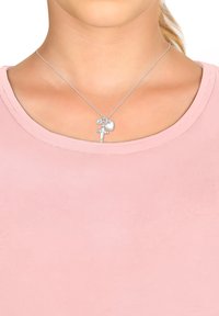 Sølv halskæde med en charm i form af et kors, et hjerte og et flag, på en tynd kæde. Båret over en lyserød, rund-halsetop.