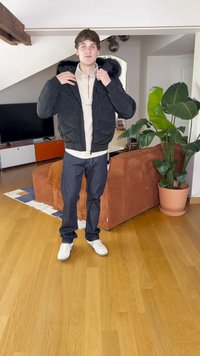 Μαύρο πλεκτό μπουφάν με γεωμετρική ραφή, με ψεύτικη γούνα στην κουκούλα, φορεμένο πάνω από ένα ανοιχτό πουκάμισο, συνδυασμένο με σκούρα τζιν και λευκά αθλητικά παπούτσια.