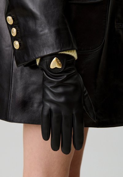Guanto in pelle nera con un'accenzione dorata a forma di cuore sul polso, caratterizzato da una texture liscia e una vestibilità su misura, indossato con un cappotto nero.