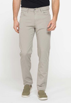 Uomo che indossa jeans beige chiaro a taglio dritto e scarpe verdi slip-on, in piedi su uno sfondo bianco semplice.