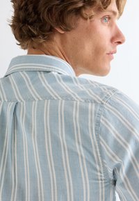 Νέος άνδρας με ανοιχτό καστανά σγουρά μαλλιά, φοράει μπλε και λευκό ριγέ πουκάμισο με γιακά, κοιτώντας πλάγια πάνω από έναν απλό φόντο.