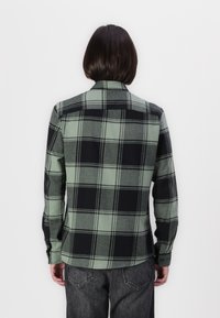 Camisa de franela a cuadros verdes y negros con cuello, botón en la parte delantera, mangas largas y dobladillo curvo. Textura de tela suave.