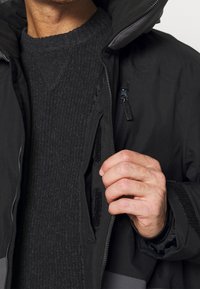 Veste imperméable noire avec des accents gris, dotée d'une fermeture éclair avant, de poches à scratch et d'un col montant, portée sur un pull noir texturé.