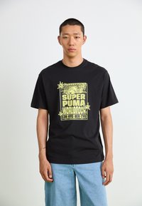 Black cotton t-shirt featuring a large yellow graphic print with "SUPER PUMA WORLD TOUR" and plant motifs, paired with light blue jeans.
