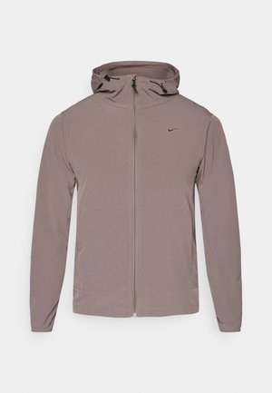 Kerge, pruun Nike jakk kapuutsiga, esiküljel tõmblukk ja kaks küljetaskut. Omab diskreetset logo rinnal ja elastseid mansette.