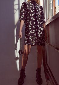 Černé květinové šaty s bílými květinovými vzory, nabíranými rukávy, volánkovým lemem, kombinované s černými kotníkovými botami a malou taškou na řetízku.