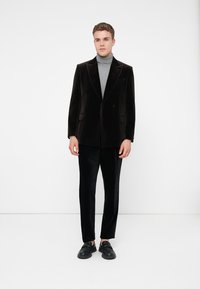 Blazer de veludo preto com lapelas pontiagudas, usado sobre um suéter de gola alta cinza. Combinado com calças de veludo preto e sapatos pretos deslizes. Design minimalista.