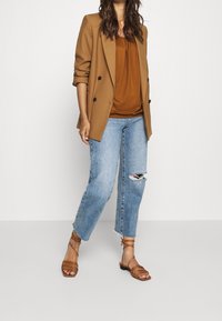 Blazer marrone doppiopetto sopra un top color ruggine, abbinato a jeans chiari strappati e sandali a tracolla aperti marroni.