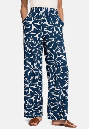 Femme portant un pantalon ample à jambes larges bleu marine avec un imprimé floral abstrait blanc, doté d'une taille élastique et de poches, associé à des sandales marron ouvertes.