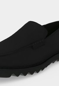 Μαύρο παπούτσι τύπου slip-on με υφασμένη επιφάνεια και οδοντωτή καουτσούκ σόλα, σχεδιασμένο για καθημερινή ή ημιεπίσημη χρήση.