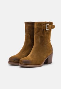 Botas de tobillo de ante marrón con tacón cuadrado, detalle de hebilla lateral, contornos lisos y puntera ligeramente desgastada, presentadas sobre un fondo blanco.