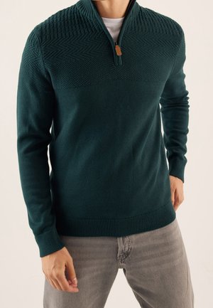 Mann trägt einen dunkelgrünen, strukturierten Strickpullover mit Halb-Reißverschluss-Kragen, kombiniert mit hellgrauer Hose, stehend vor einfachem Hintergrund.