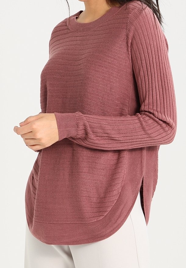 Kobieta ubrana w sweter z prążkowanej dzianiny w kolorze jasnoróżowym, z długimi rękawami i bocznymi rozcięciami, zestawiony z jasnymi spodniami.
