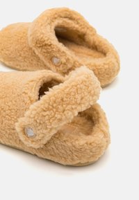 Zachte, donzige beige slippers met verstelbare banden en een klein rond knoopdetail op een witte achtergrond.