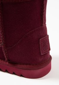 Botín de ante burdeos hasta el tobillo con una superficie texturizada, costura reforzada y suela de goma con un patrón de banda de rodadura en zigzag; parche etiquetado en el costado.