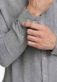 Primo piano di una camicia in lino grigia, con polsini abbottonati, bottoni chiari a contrasto e una sottile trama del tessuto. Mano che regola il polsino.