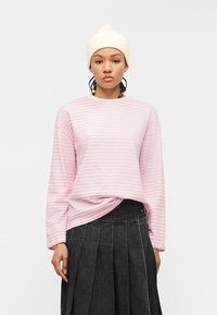 Νεαρή γυναίκα που φοράει λευκό πλεκτό σκουφάκι, ροζ και λευκή ριγέ μπλούζα με μακριά μανίκια και σκούρο πλισέ φούστα, κοιτάζοντας μπροστά με ουδέτερη έκφραση.