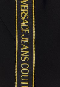 Tissu noir avec une étiquette tissée verticale noire et jaune portant la mention « VERSACE JEANS COUTURE » en lettres grasses.