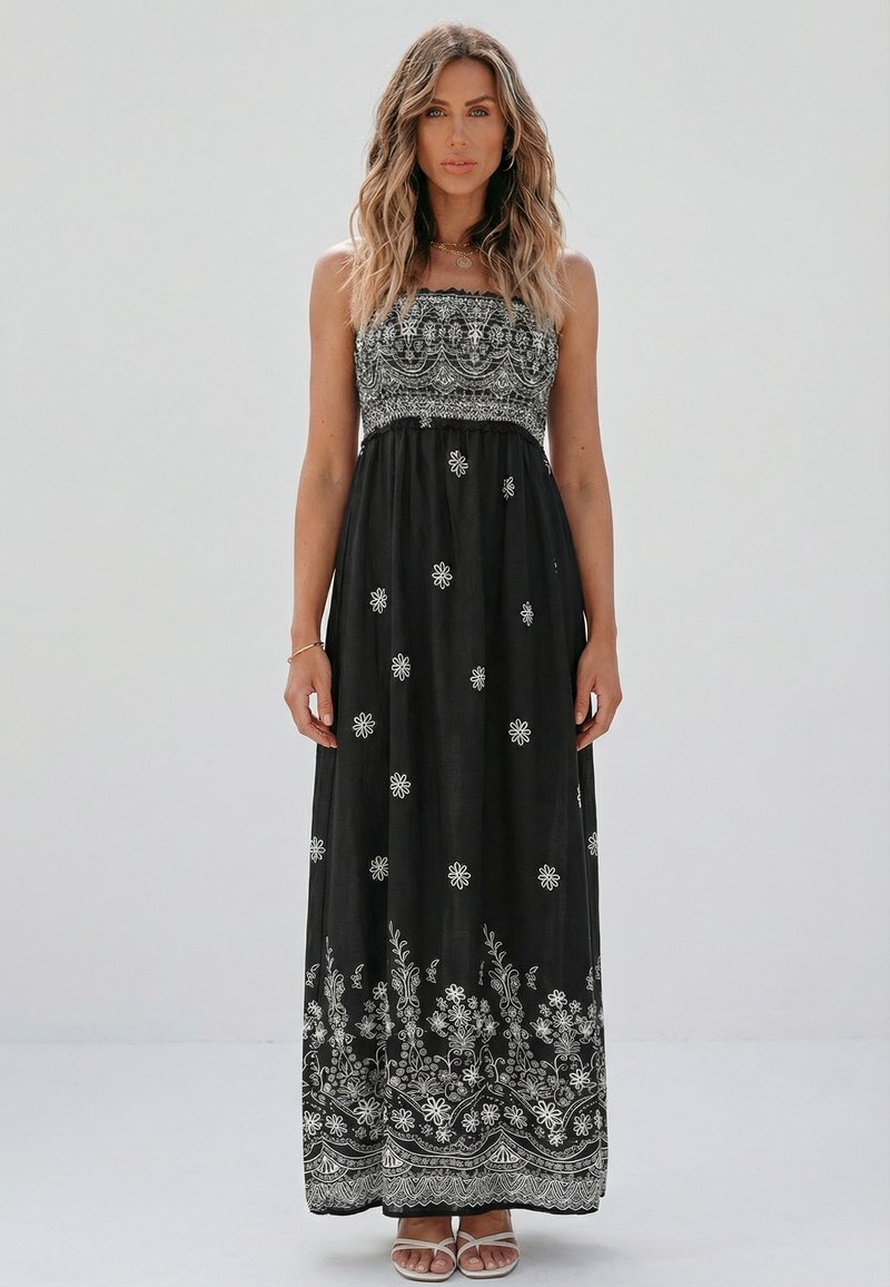 Donna con capelli mossi che indossa un abito lungo nero senza spalline con ricami floreali bianchi, in piedi contro uno sfondo chiaro e uniforme.