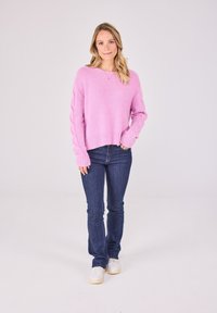 Helles rosa gestricktes Sweatshirt mit lockerer Passform und geripptem Saum, kombiniert mit dunkelblauen Jeans und weißen Sneakern. Glatte Textur mit einem gemütlichen Erscheinungsbild.